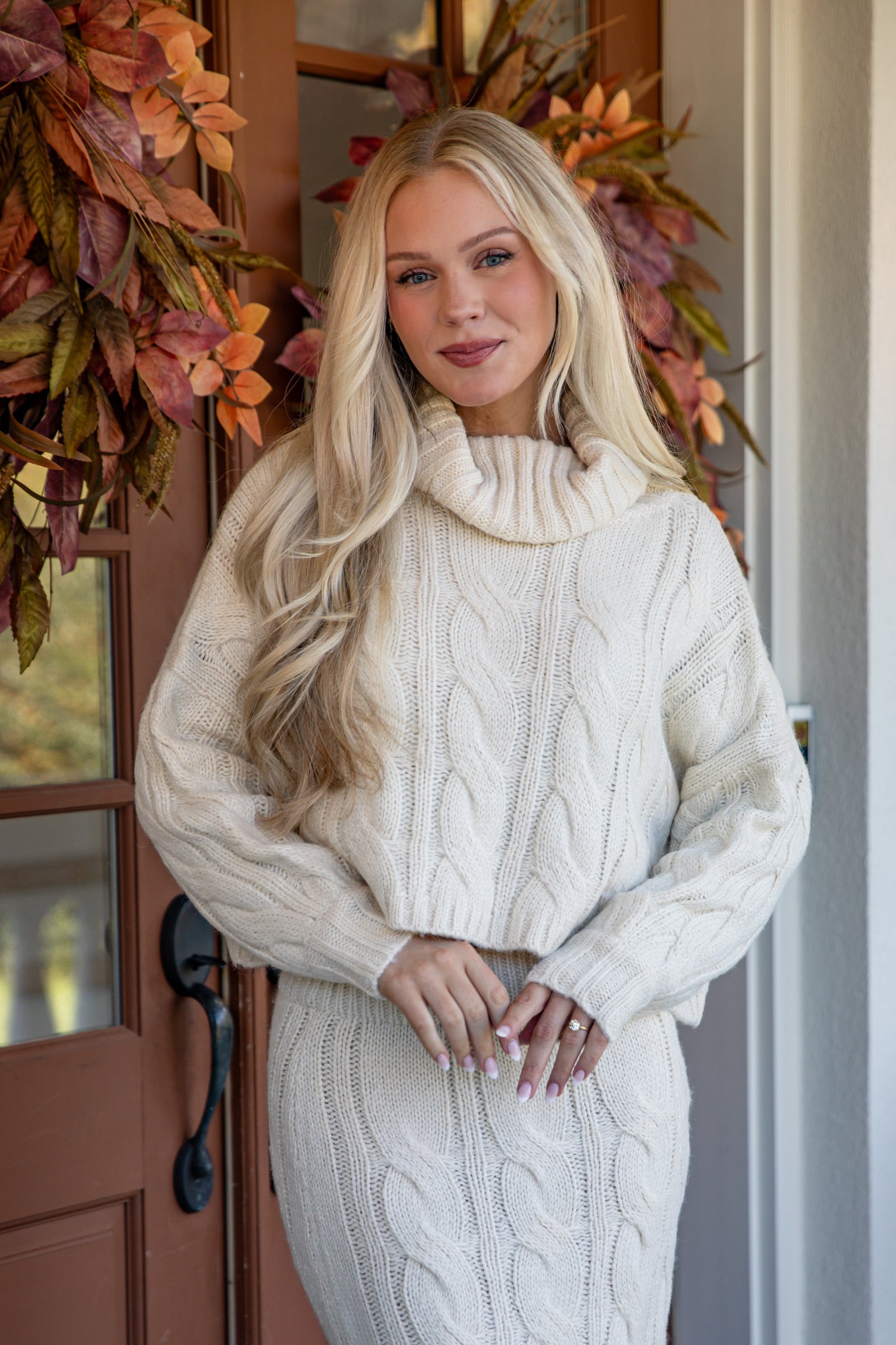 Woman wearing a cream cable knit sweater in front of a door with autumn decorations.