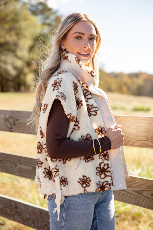 Woman wearing a white floral-patterned vest outdoors with a wooden fence and trees in the background.