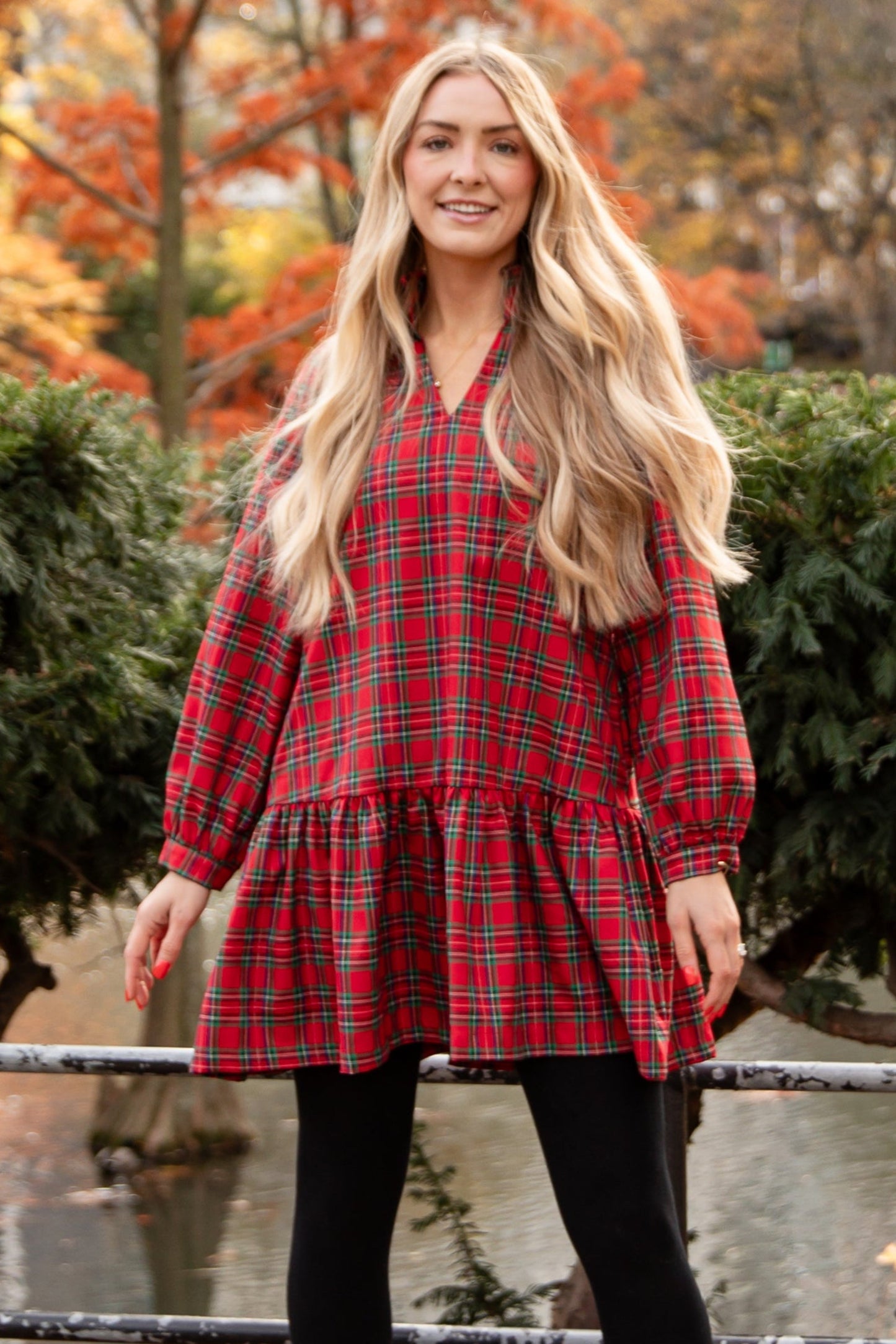 Woman in a red plaid dress standing in an urban park with autumn foliage.