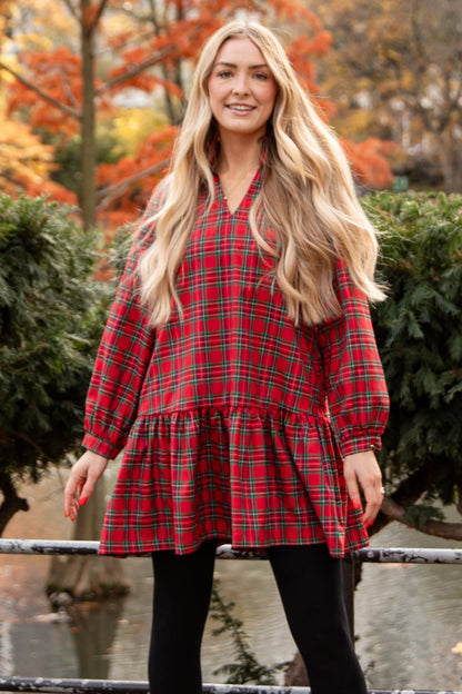 Woman in a red plaid dress standing in an urban park with autumn foliage.