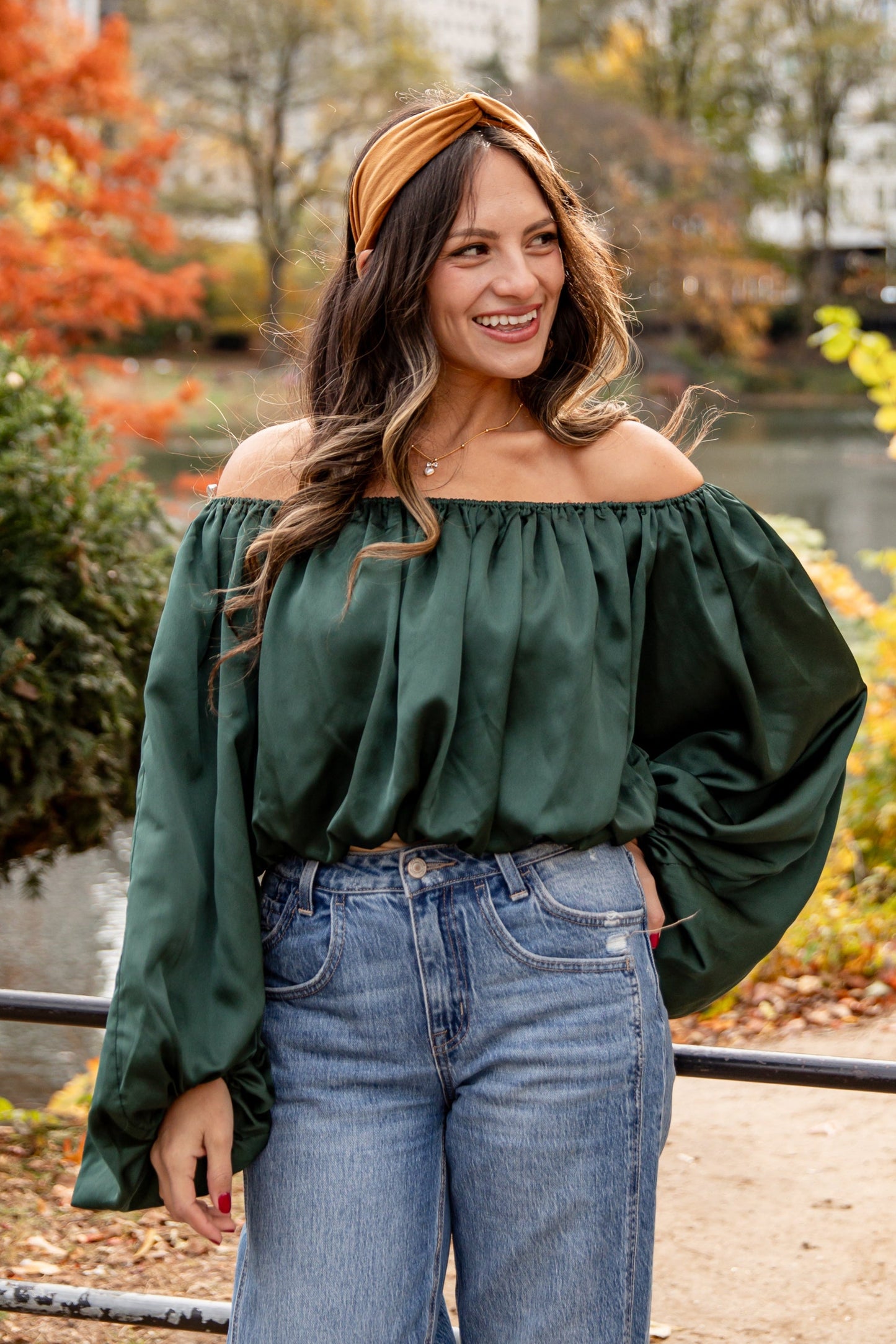 Woman in a green top and blue jeans standing in an urban park with trees and buildings in the background.
