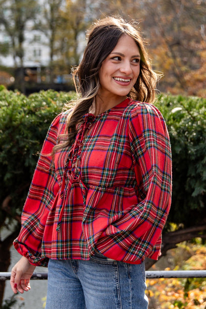 Woman wearing a red plaid shirt and jeans standing in an urban park with trees and buildings in the background.