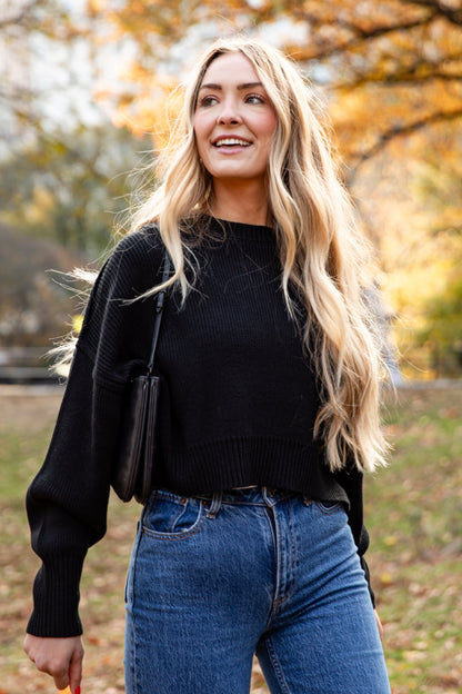 Woman in black sweater and blue jeans standing in a park with autumn trees.