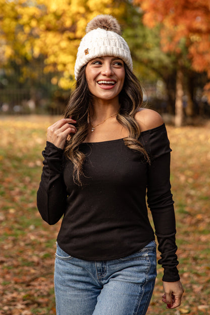 Woman wearing a black long-sleeve top, blue jeans, and a white knit beanie with a pom-pom in an autumn setting.