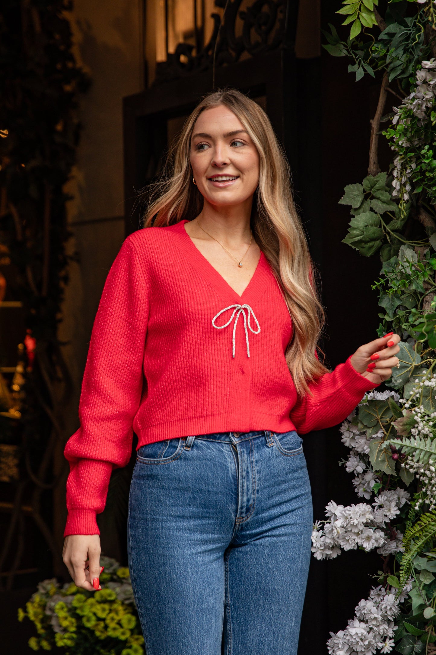 Woman wearing a red sweater with a white bow design and blue jeans, standing outdoors with plants in the background.