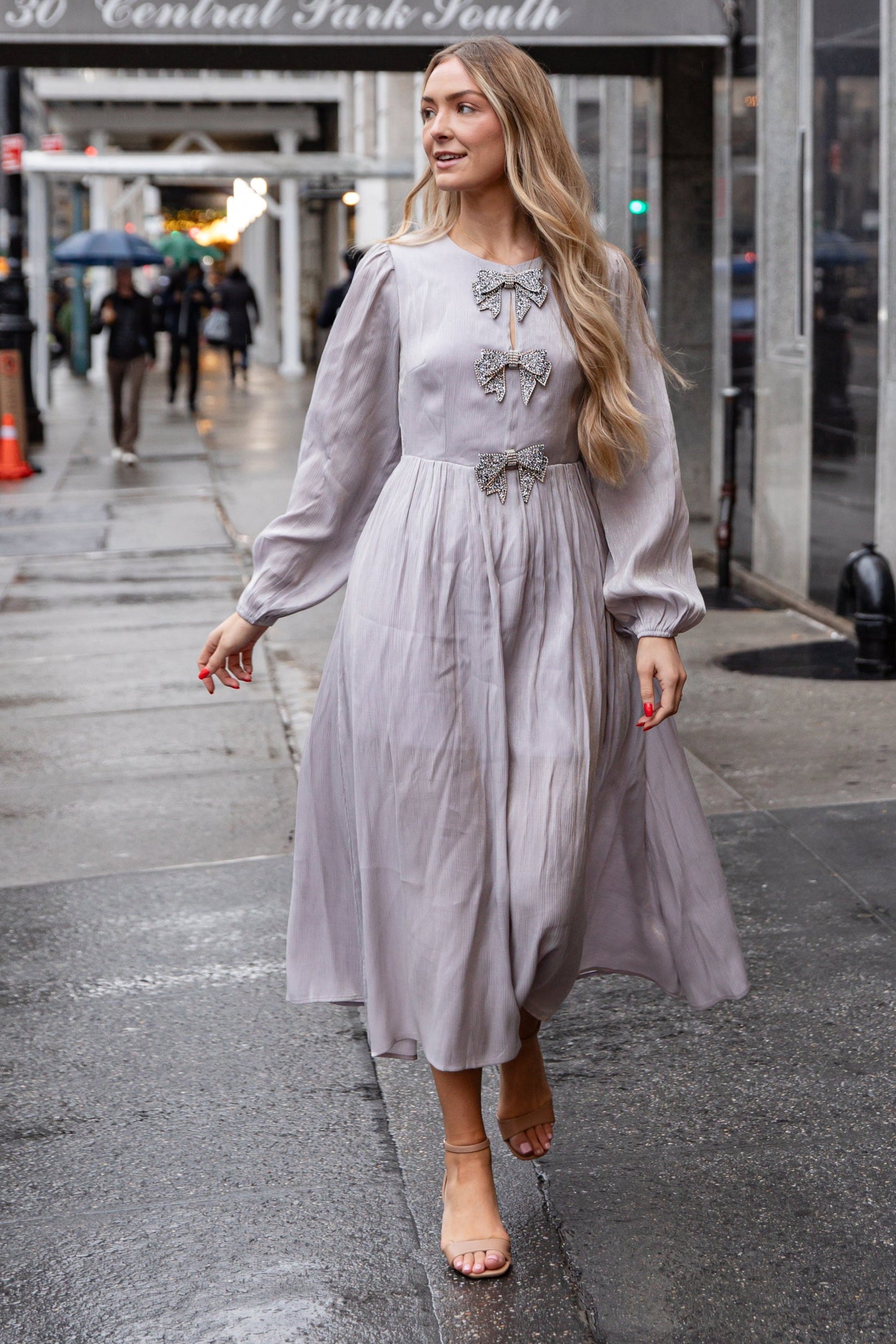 Woman in a light gray dress walking on a city street with '30 Central Park South' in the background.