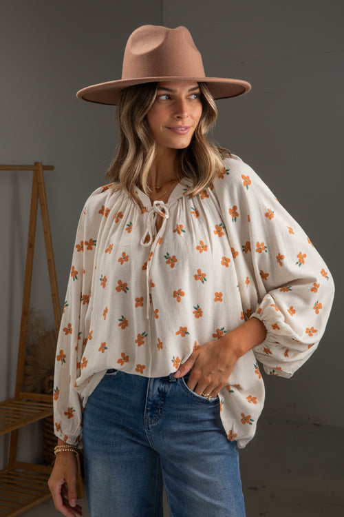 Woman wearing a floral blouse, jeans, and a brown hat against a plain background