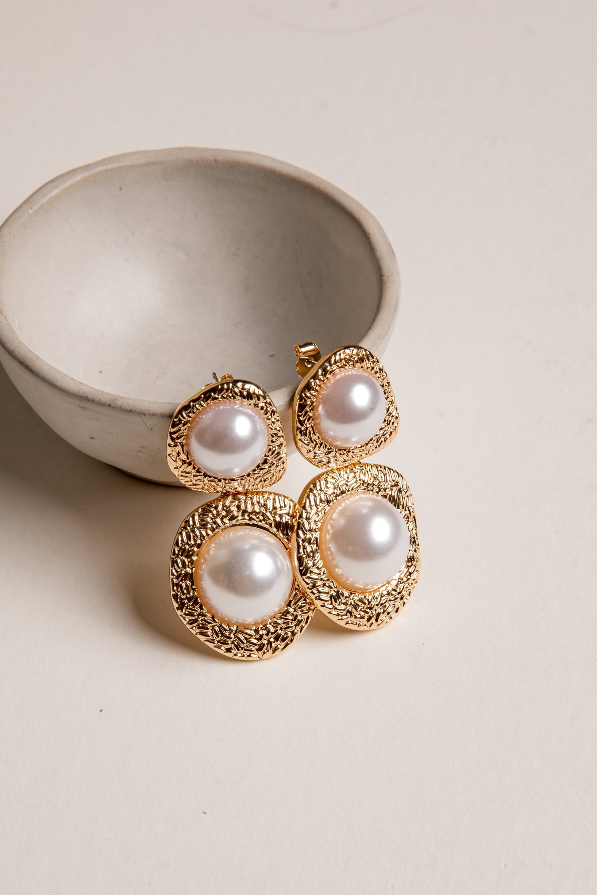 Gold-framed pearl earrings on a neutral background